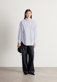 Svetlomodrá košeľa na zapínanie s náprsným vreckom, v kombinácii s tmavými voľne strihanými nohavicami a čiernou kabelkou na neutrálom pozadí.