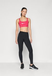 Ružičasti sportski grudnjak s bijelim Nike logotipom, u kombinaciji s crnim tajicama visokog struka. Model nosi crne sportske tenisice s bijelim detaljima.