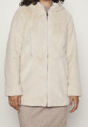 Personne portant une veste à capuche zippée en fausse fourrure crème par-dessus une robe en tricot beige, visible du cou jusqu'à mi-cuisse.