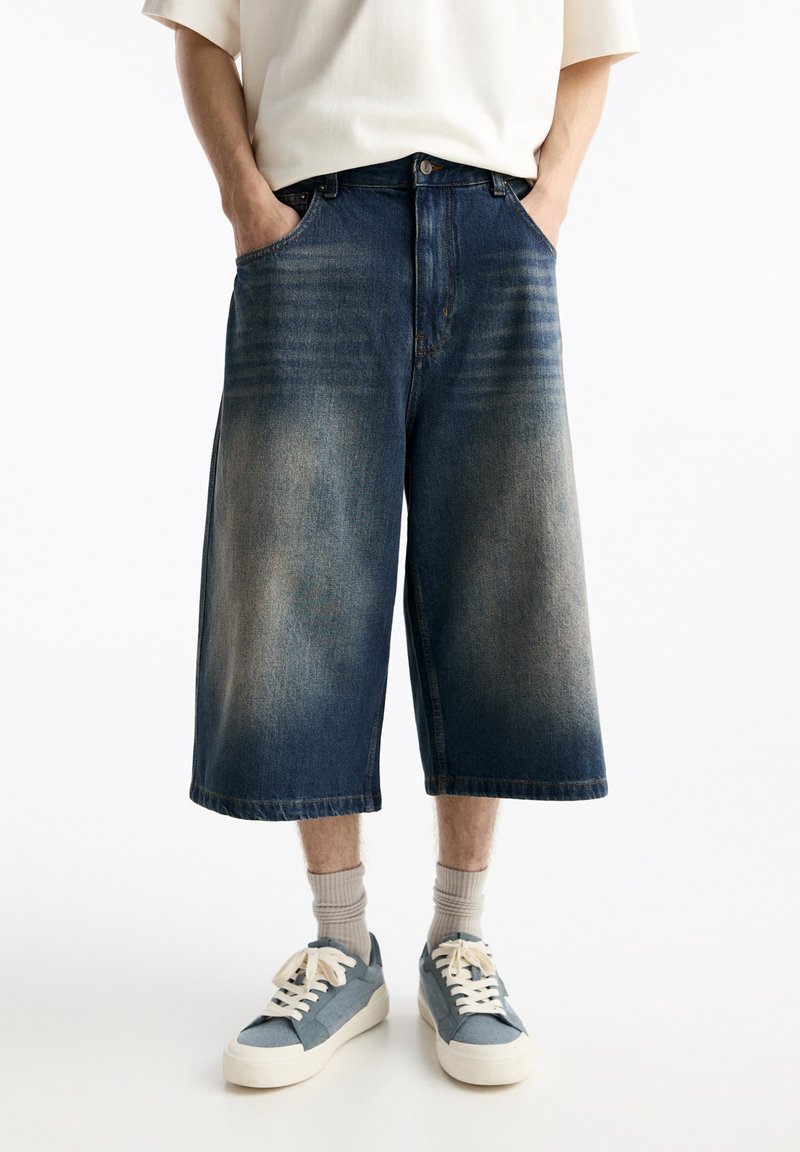 Personne portant un short en jean bleu délavé à jambes larges, des chaussettes beige clair et des baskets bleu et blanc, debout les mains dans les poches.