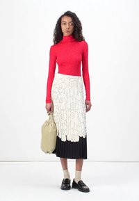 Rotes Langarm-Oberteil mit floralen Muster, kombiniert mit einem plissierten Midirock, der oben weiß und unten schwarz ist. Das Modell hält eine weiche beige Tasche.