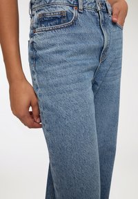 Jasnoniebieskie dżinsy z dopasowaną talią, prostym krojem nogawek, widocznym przeszyciem i miękką teksturą. Podkreślone detale kieszeni.