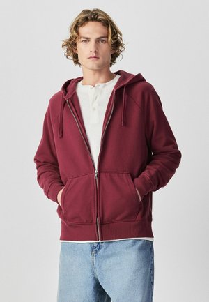 Young man with curly hair wearing a zipped maroon hoodie over a white henley shirt and light blue jeans, hands in hoodie pockets.