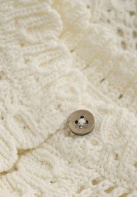 Cremefarbener Strickstoff mit einem strukturierten Muster, der einen runden, braunen Knopf mit vier Löchern zeigt. Die Nahaufnahme betont die Weichheit und die Details.