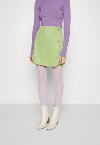 Saia verde envolvente com dois botões, combinada com meias-calças lilás e botas brancas de tornozelo. O tecido é suave e apresenta um design estruturado.