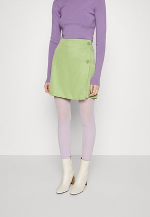 Saia verde envolvente com dois botões, combinada com meias-calças lilás e botas brancas de tornozelo. O tecido é suave e apresenta um design estruturado.
