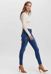 Top de manga larga crema combinado con vaqueros ajustados de color azul oscuro y tacones altos marrones, mostrando una silueta ajustada y una textura suave.