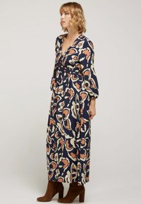 Robe maxi en bleu marine foncé avec des motifs abstraits beige et rouille, dotée d'un décolleté en V profond, de longues manches et d'une taille cintrée, accompagnée de bottes à talons marron.