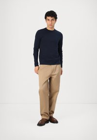 Marineblaues Strickpullover mit Rundhalsausschnitt, kombiniert mit hellbeigen Hosen. Braune Loafers runden das Outfit ab. Glatte Textur, lässige Passform.