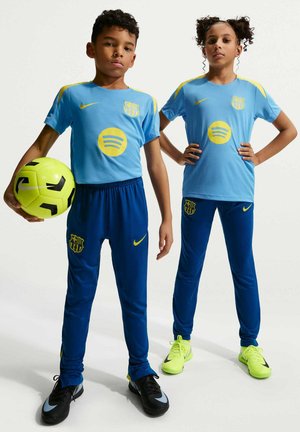 Dwoje dzieci w niebiesko-żółtych strojach piłkarskich FC Barcelona, jedno trzyma neonowo-żółtą i czarną piłkę, stoją pewnie na jednolitym tle.