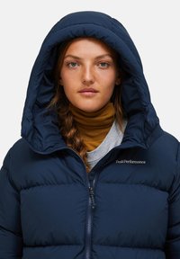 Navyblauer Pufferjacke mit großer Kapuze, gestepptem Material und Frontreißverschluss; trägt ein Logo auf der Brust und wird über einem Rollkragenpullover getragen.