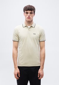 Jeune homme aux cheveux bouclés portant un polo beige avec un bord noir et le logo "BOSS", debout devant un fond clair uni.