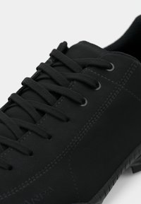 Black sneaker made of smooth synthetic material, featuring a rounded toe, flat laces, and subtle textured accents on the sole.