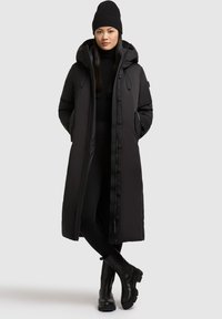 Dlouhý černý péřový kabát s kapucí, zipy a bočními kapsami, nošený přes černý svetr a legíny, doplněný černými kotníkovými botami.