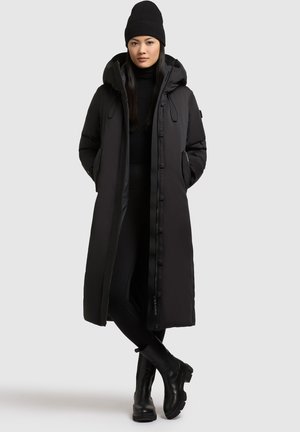 Langer schwarzer Puffercoat mit Kapuze, Reißverschlüssen und Seitentaschen, getragen über einem schwarzen Pullover und Leggings, kombiniert mit schwarzen Stiefeletten.