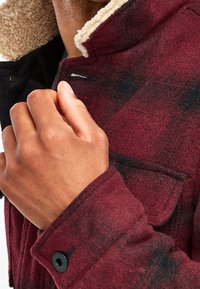 Rood en zwart geblokt jack met een fleece kraag, voorzien van knoopaccenten, een textuurstof en een zichtbare borstzak in de afbeelding.