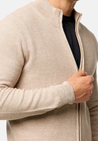 Hombre vestido con un suéter de cremallera texturizado beige sobre una camisa negra, sosteniendo la cremallera a medio bajar cerca del pecho.