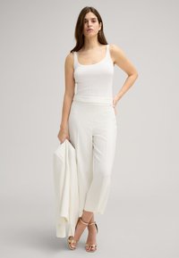 Canotta bianca a costine abbinata a pantaloni bianchi su misura. Il modello tiene in mano un blazer bianco e indossa sandali con cinturini dorati. Design semplice, texture liscia.