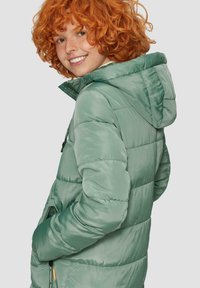 Veste matelassée verte avec capuche, design matelassé, poches latérales et texture lisse. Le modèle a des cheveux bouclés orange.