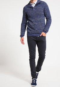 Strickpullover in Navy mit Knopfkrause, kombiniert mit schmal geschnittenen dunklen Hosen und navyfarbenen Sneakers, die einen lässigen, gestuften Look kreieren.
