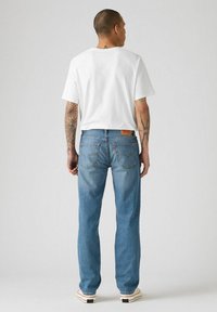 Vista trasera de un hombre vistiendo una camiseta blanca de manga corta, jeans de mezclilla azul claro con costuras y marcas visibles, y zapatillas de color crema.