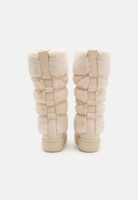 Beige fløyelsknelange støvler med teksturert overflate, som har elastiske detaljer langs sidene og en glatt gummisåle.