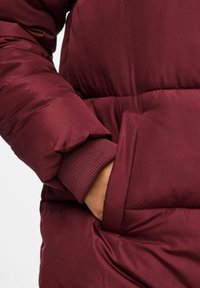 Bordo pūkinis švarkas su lygiu paviršiumi, su raukšlėta apykakle, šoninėmis kišenėmis ir siuvinėjimu. Parodytas riešo detalės priartinimas.