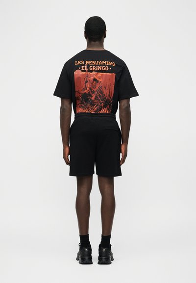 Άνδρας που φοράει μαύρο σορτς και μαύρο T-shirt με πορτοκαλί γραφικό και κείμενο «LES BENJAMINS EL GRINGO» στην πλάτη, με την πλάτη γυρισμένη.