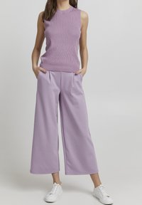 Haut sans manches en rib ou dans un violet clair associé à un pantalon ample violet clair. Le mannequin porte des baskets blanches, les mains dans les poches. Texture lisse.