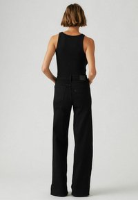 Jean noir à jambes larges en denim, avec une taille haute et deux poches arrière. Le modèle porte un débardeur noir, vu de dos.