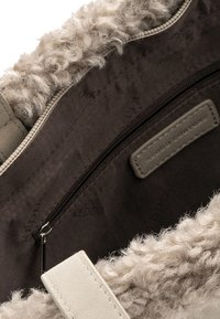 Detaljan prikaz otvorene torbe ukrašene bež umjetnim krznom, s crnim tkaninskim unutrašnjim dijelom, džepom na patentni zatvarač i kožnom etiketu s natpisom "Keddo Couture".