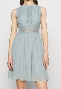 Ljusblå klänning med en spetslivet som har cirkulära mönster, ärmlös design och en svävande kjol. Texturerad spets ger detalj vid midjan.