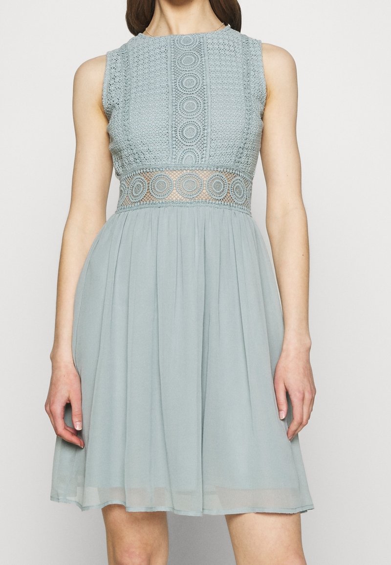 Ljusblå klänning med en spetslivet som har cirkulära mönster, ärmlös design och en svävande kjol. Texturerad spets ger detalj vid midjan.