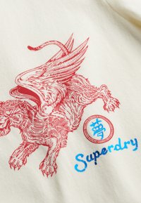 Witte stof met een rood beeld van een gevleugelde leeuw en een ronde insigne met blauwe tekst "Superdry." Details van de textured print zijn zichtbaar.