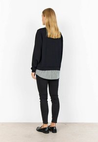 Frau mit blonden Haaren, die einen schwarzen Pullover über einem gestreiften Shirt, schwarze Skinny Jeans und schwarze Slipper trägt, steht gegen eine weiße Wand.