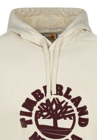 Felpa color crema con cappuccio, caratterizzata da un logo Timberland viola con grafica ad albero. Include un cappuccio con coulisse e un tessuto testurizzato.