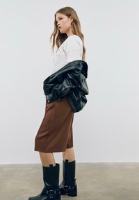 Giacca di pelle nera appoggiata su una camicia bianca, abbinata a pantaloni corti marroni fino al ginocchio e stivali neri alti fino al ginocchio con fibbie.