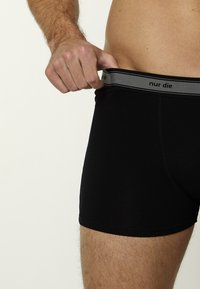 Czarne bawełniane bokserki z szarą elastyczną talią z napisem "nur die", pokazane podczas ściągania przez rękę.