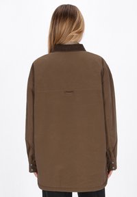 Brun oversized jakke med et kordfløjlskrage, synlige sømme og knaplukninger ved ærmerne, lavet af et glat og slidstærkt stof.