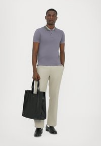 Camisa polo morada con cuello a rayas, pantalones beiges y un bolso negro. Modelo vistiendo zapatos negros, de pie frente a un fondo blanco.