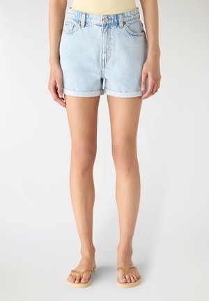 Donna con pantaloncini di jeans azzurri con orlo arrotolato e infradito beige, in piedi davanti a uno sfondo semplice.