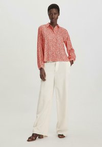 Camicia rossa a maniche lunghe con colletto, abbinata a pantaloni bianchi a gamba larga e sandali con tacco impilato color marrone.