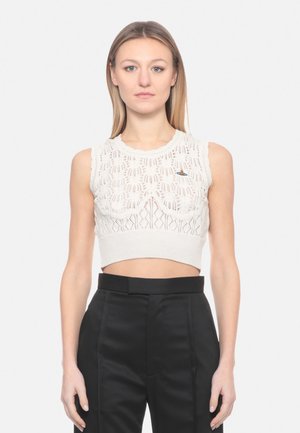 Giovane donna che indossa un top corto a maglia bianco senza maniche con dettagli a motivo e pantaloni neri a vita alta, in piedi contro uno sfondo semplice.