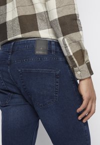 Jeans de mezclilla azul oscuro con corte recto, que presentan un parche de cuero negro con la marca en la cinturilla y un detalle en el bolsillo trasero.