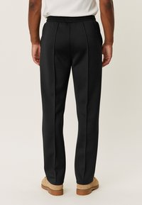 Pantalons noirs ajustés avec une texture lisse, dotés d'une taille à cordon de serrage et de plis sur les côtés. Portés avec des chaussures beiges.
