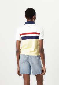 Polo-shirt med gul krop, hvide ærmer og vandrette striber i rød og marineblå. Har en marineblå krave og en v-formet bagsøm.