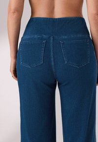 Pantaloni din denim albastru cu talie înaltă. Caracteristicile includ două buzunare pe spate și o croială relaxată. Materialul are un aspect moale, texturat.