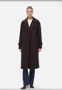 Cappotto marrone doppiopetto con rever, dotato di maniche con bottoni e vestibilità rilassata, indossato sopra un top chiaro e jeans.