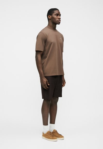 Άνδρας που στέκεται με καφέ μπλουζάκι, σκούρο καφέ σορτς, λευκές κάλτσες και ταμπά αθλητικά παπούτσια πάνω σε λευκό φόντο.