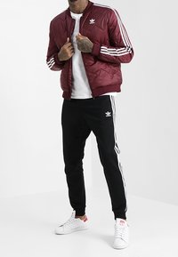 Man som bär en vinröd Adidas vadderad jacka, svarta Adidas träningsbyxor med vita ränder, vita sneakers och en vit T-shirt.